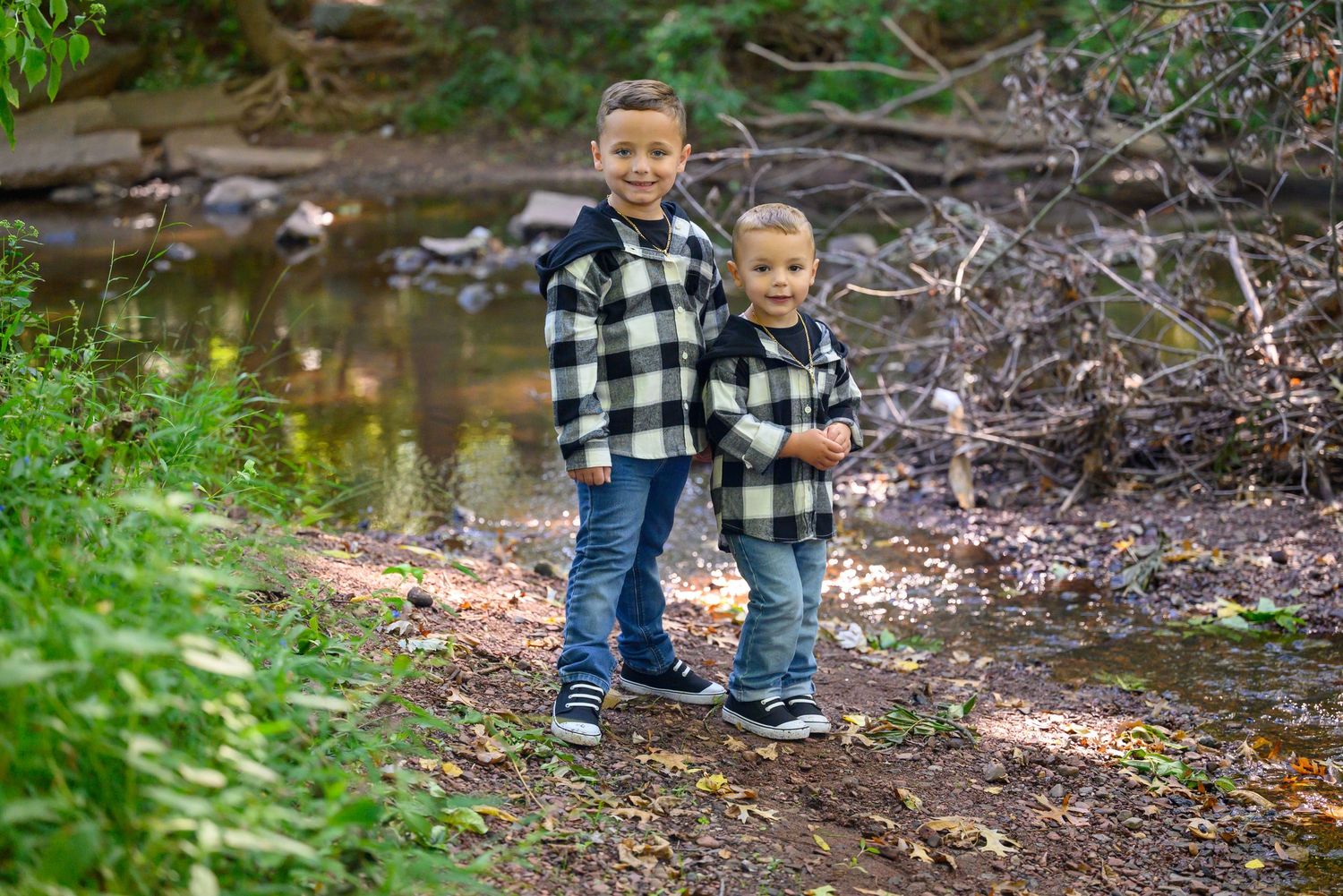 Children wearing matching plaid shirts stand together on a wooded trail near a stream in nature.