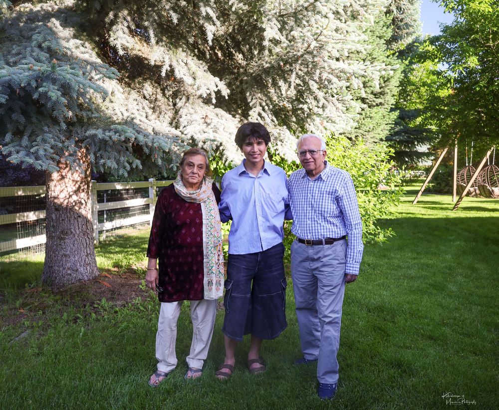 Three people standing together on a grassy lawn with trees and a wooden fence in the background on a sunny day.