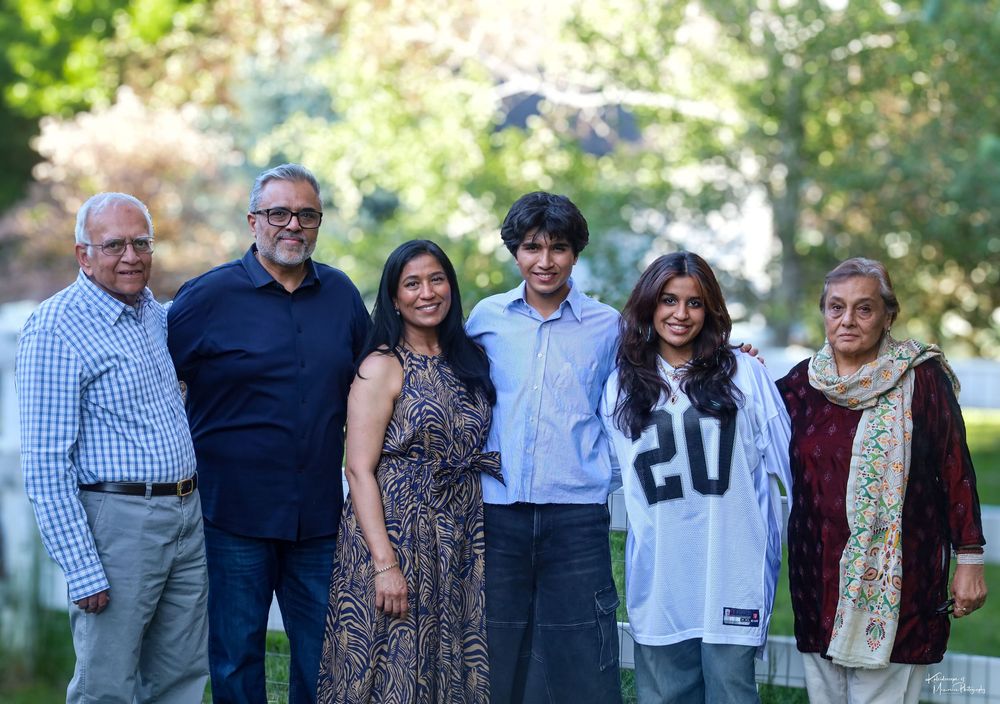 A multi-generational family poses together outdoors in a garden setting with greenery in the background.