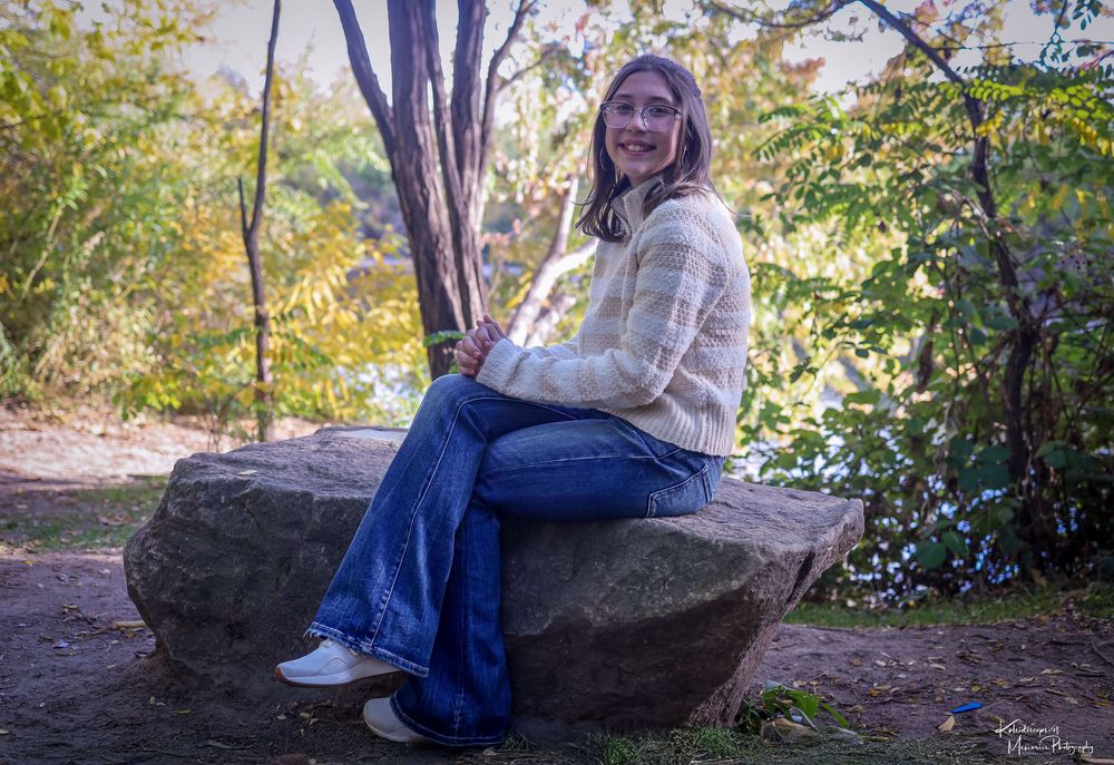 Person in beige sweater and jeans sits on large rock along wooded nature trail on sunny autumn day.