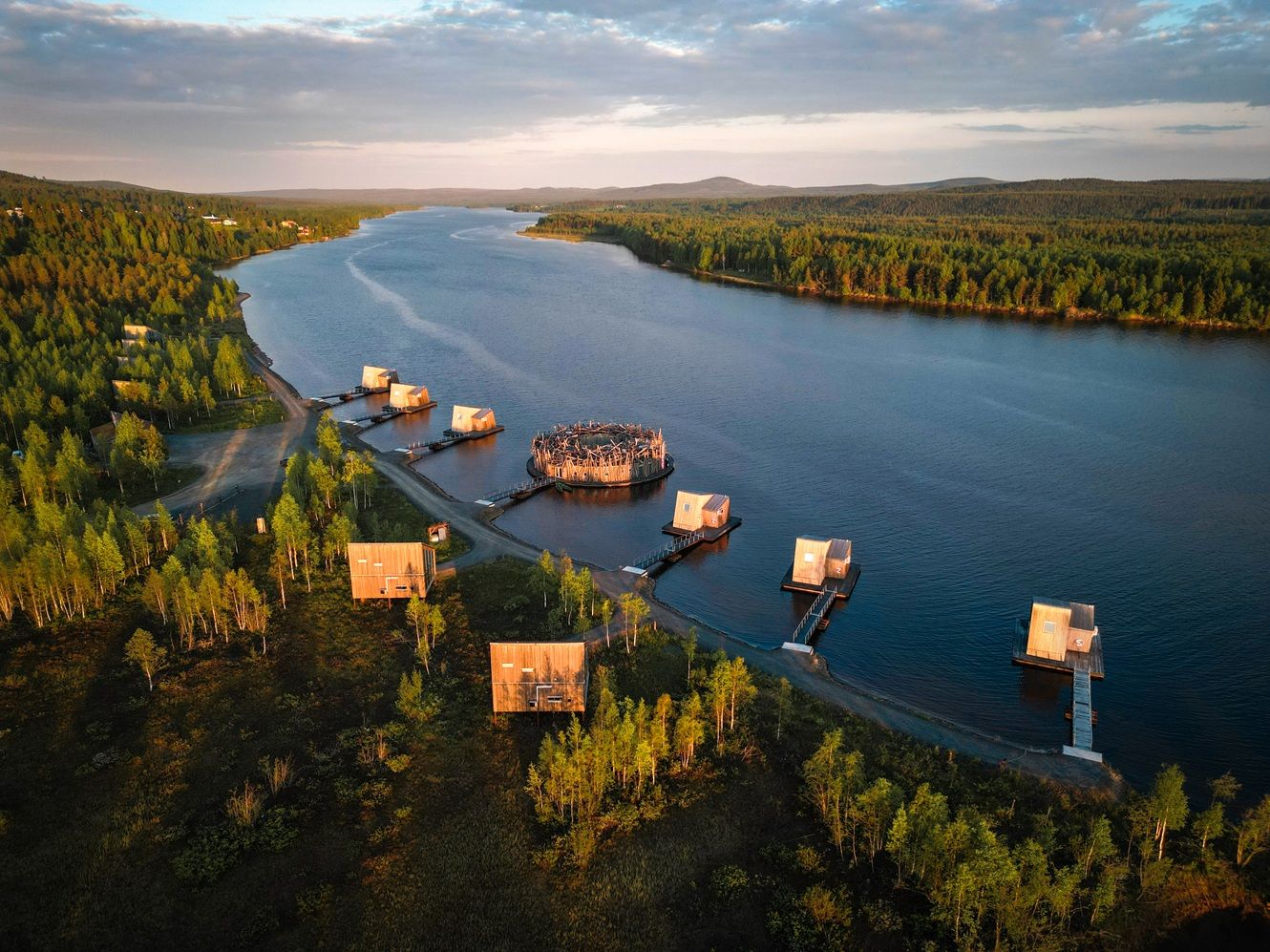 Aerial view of luxury floating cabins on a serene river surrounded by lush forests at golden hour in Lapland.
