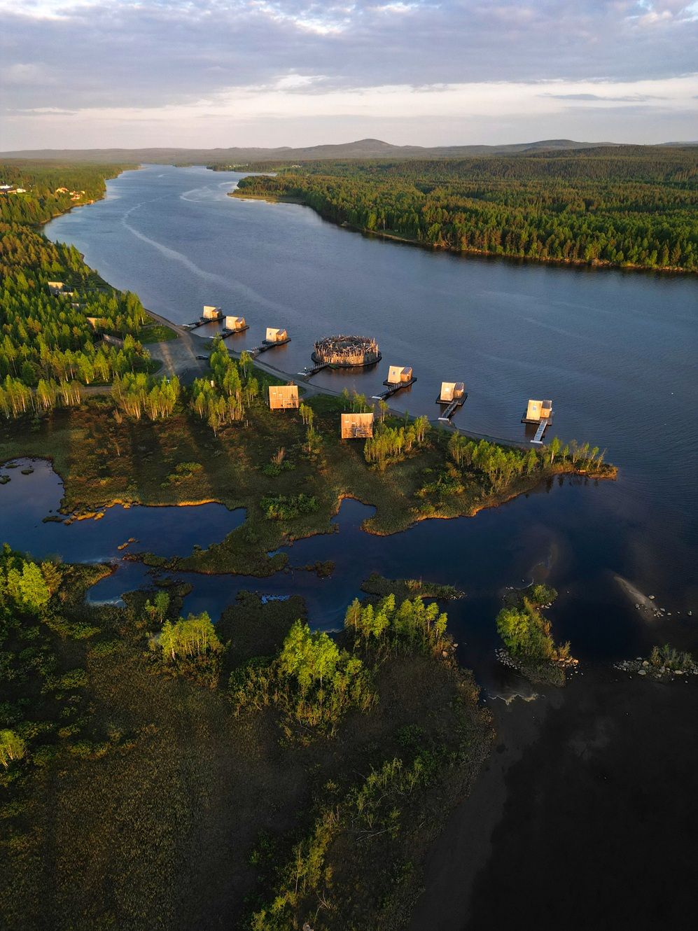Aerial view of floating cabins on a serene northern lake surrounded by lush boreal forest at golden hour.