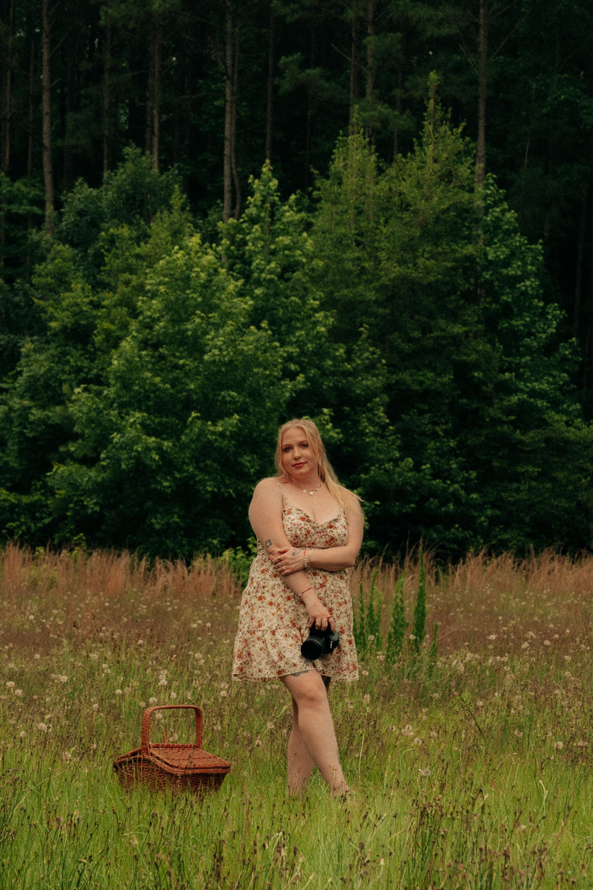 Person in teal top and black shorts holds camera while standing in wildflower meadow at sunset.