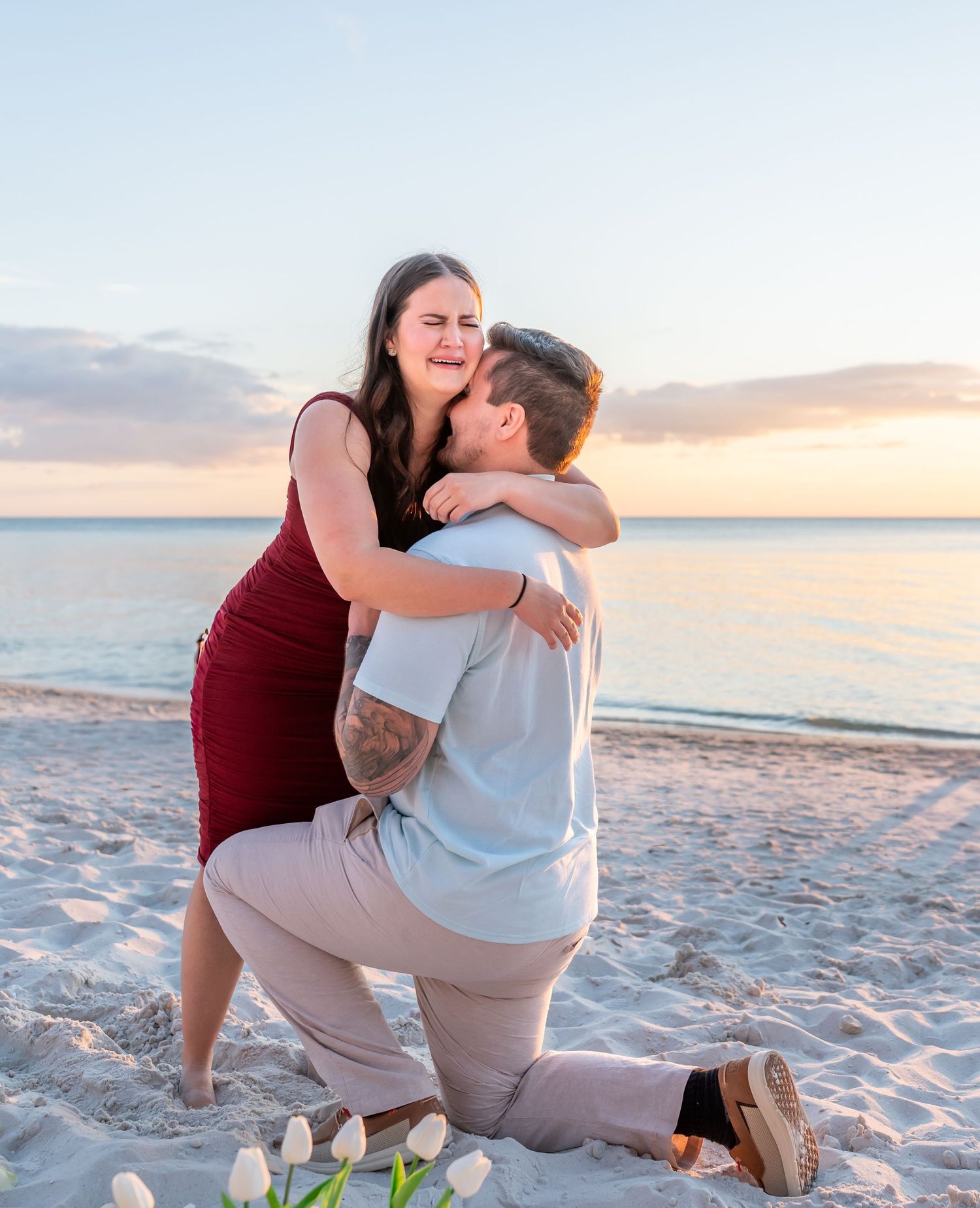 A romantic couple embraces on a sandy beach at sunset with white flowers laying nearby on the sand.