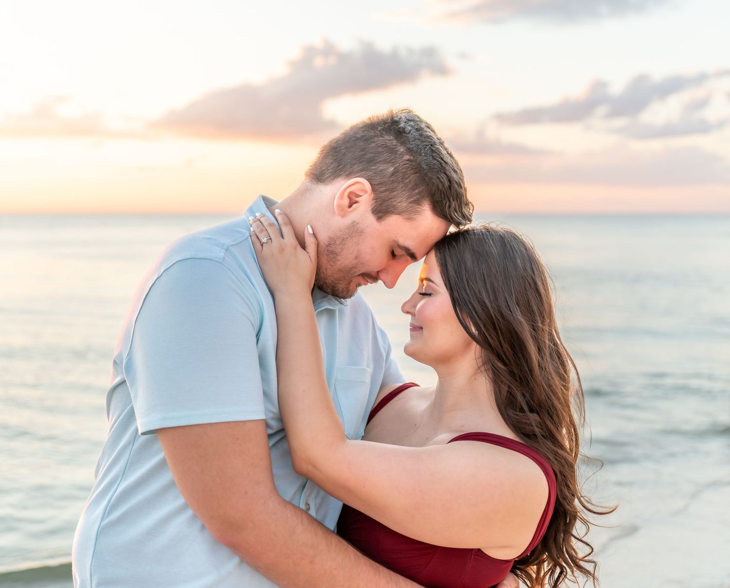 A romantic couple embraces and kisses during a sunset photoshoot on a beach with ocean waves in the background.