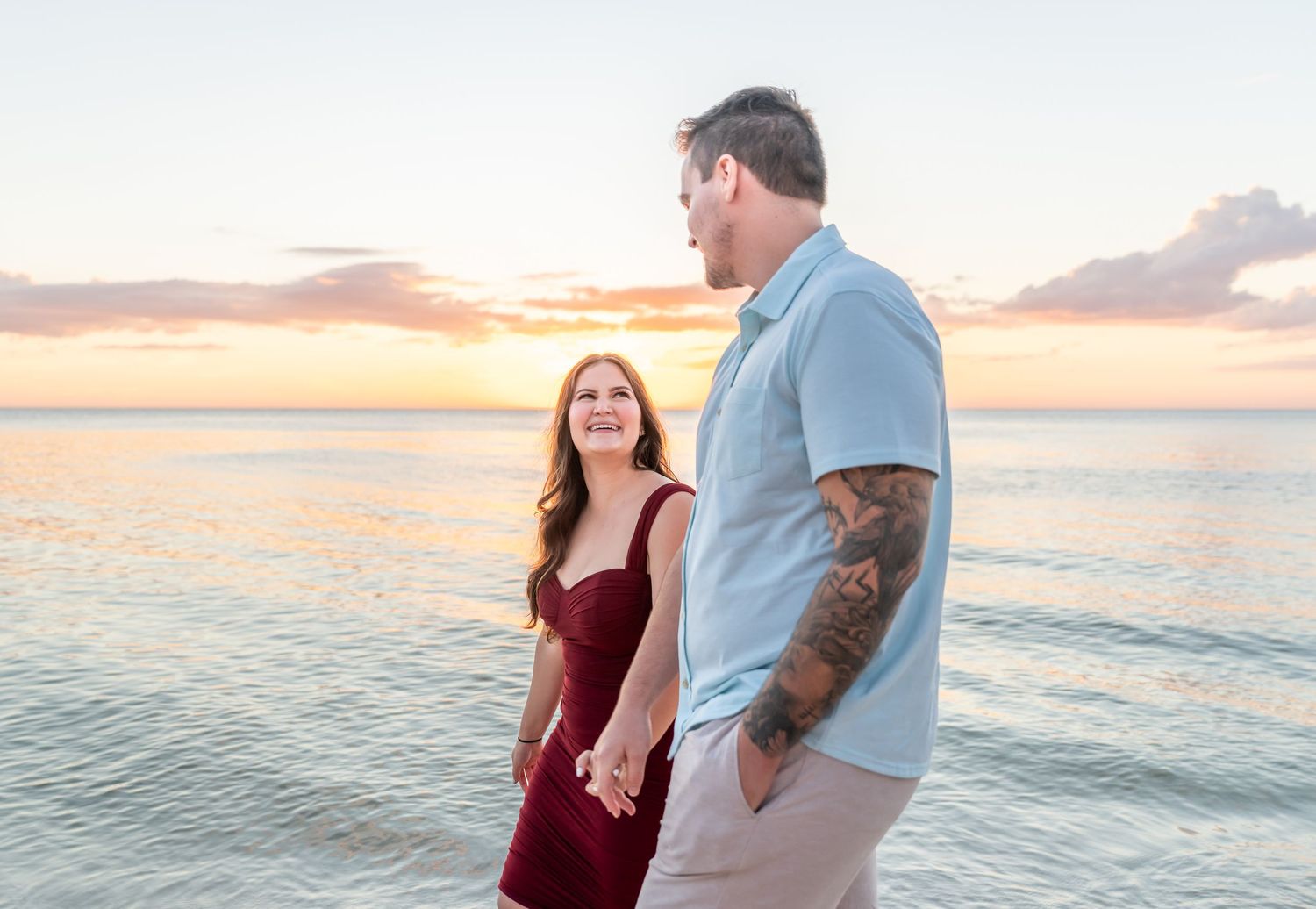 A couple stands together on a beach at sunset with the ocean waves gently rolling in the background.