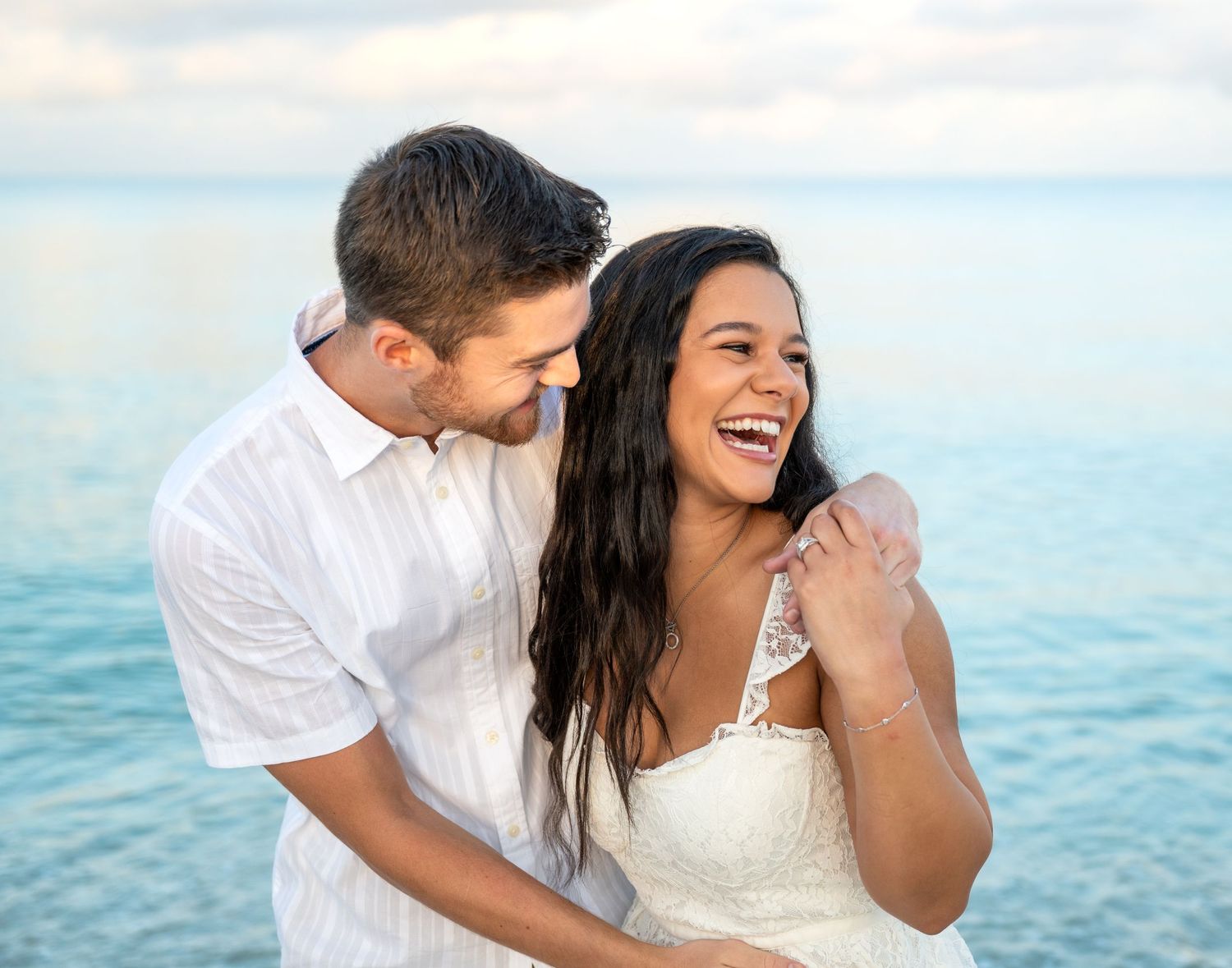 A happy couple shares joyful moments and embraces on a beach with turquoise ocean waters in the background.