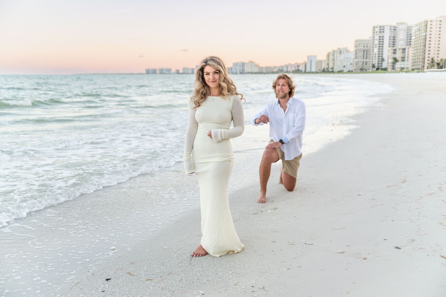 Couple in white attire walking and laughing together along a sunny beach with waves and buildings in background.