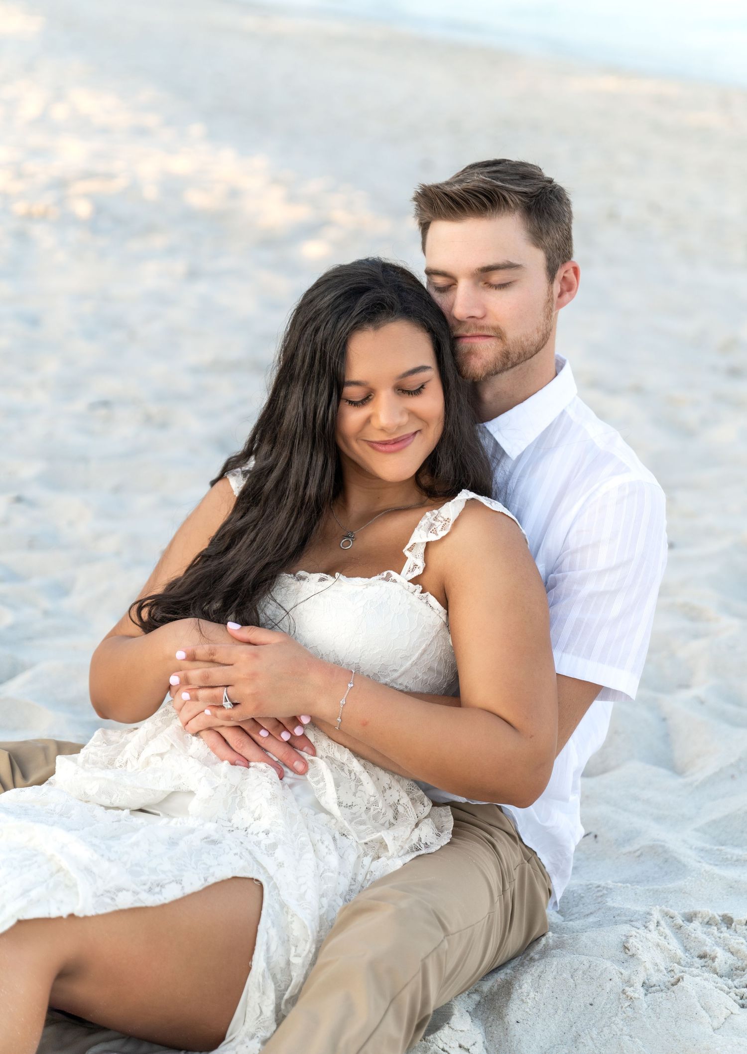 A romantic couple embraces on a sandy beach at sunset, with a woman in a white lace dress and her partner in white shirt.