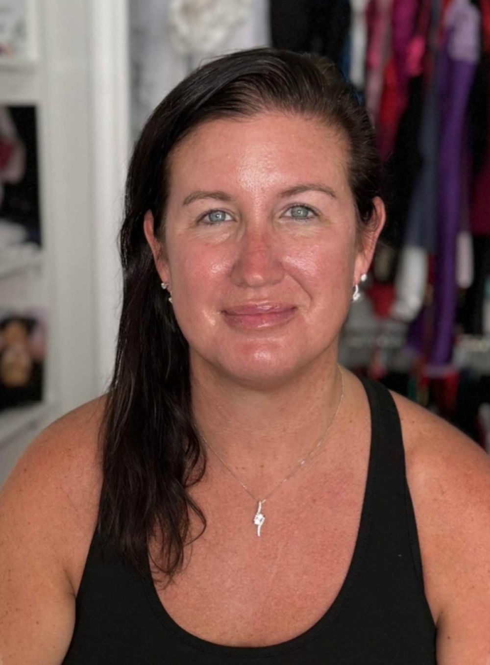 Woman with dark hair and blue eyes wearing a black tank top and necklace, smiling in a bright room.