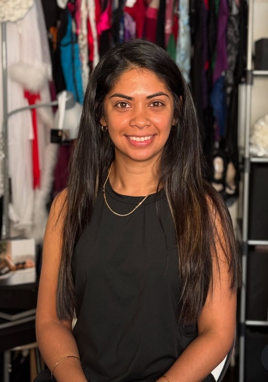 A smiling woman with long dark hair wearing a black sleeveless top and gold necklace stands in front of a clothing rack.