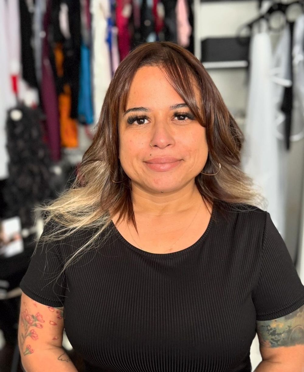 Woman with brown ombre hair and tattoos wearing a black ribbed t-shirt standing in front of a clothing rack.