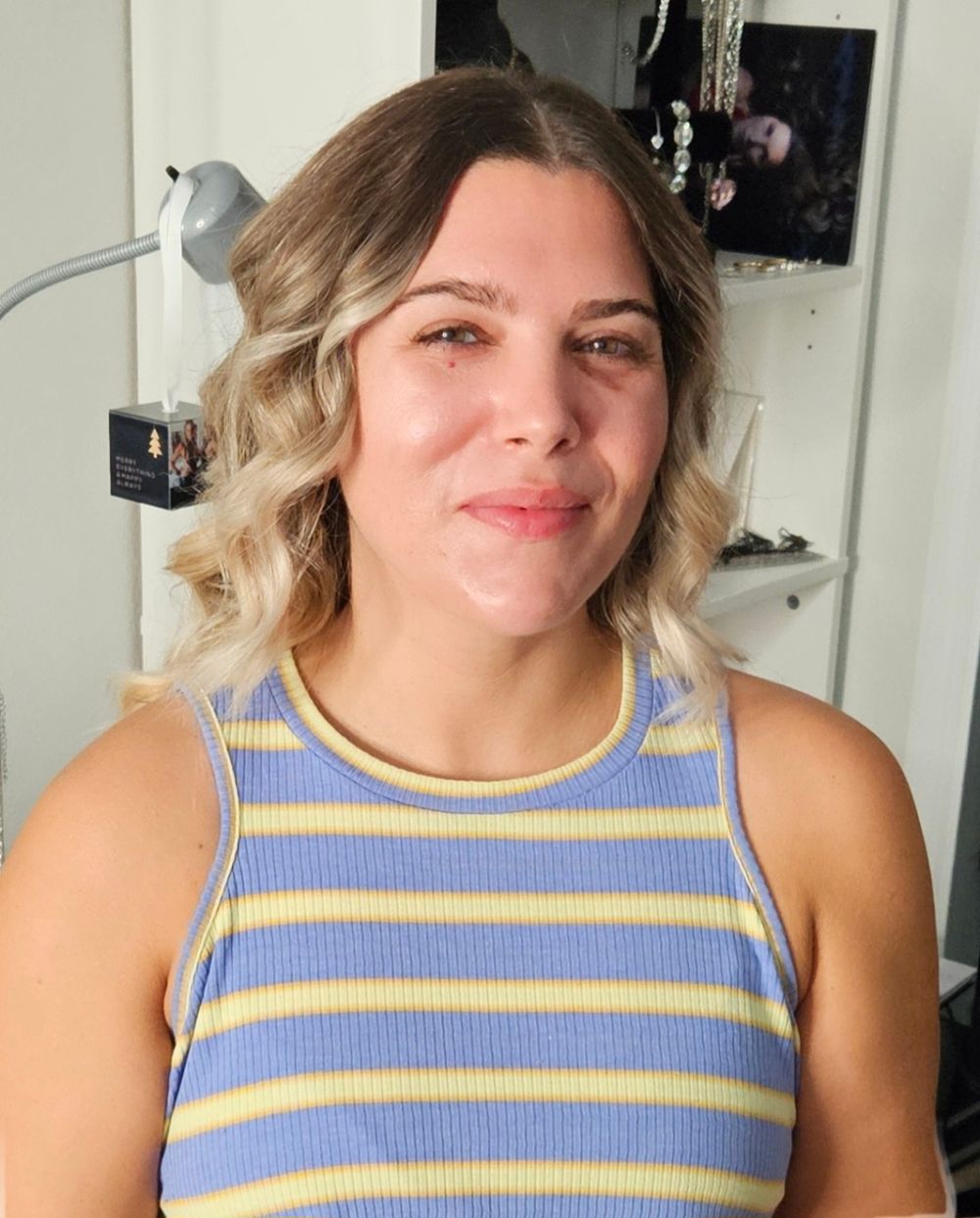 Young woman with blonde wavy hair wearing a blue and yellow striped sleeveless top, smiling indoors.