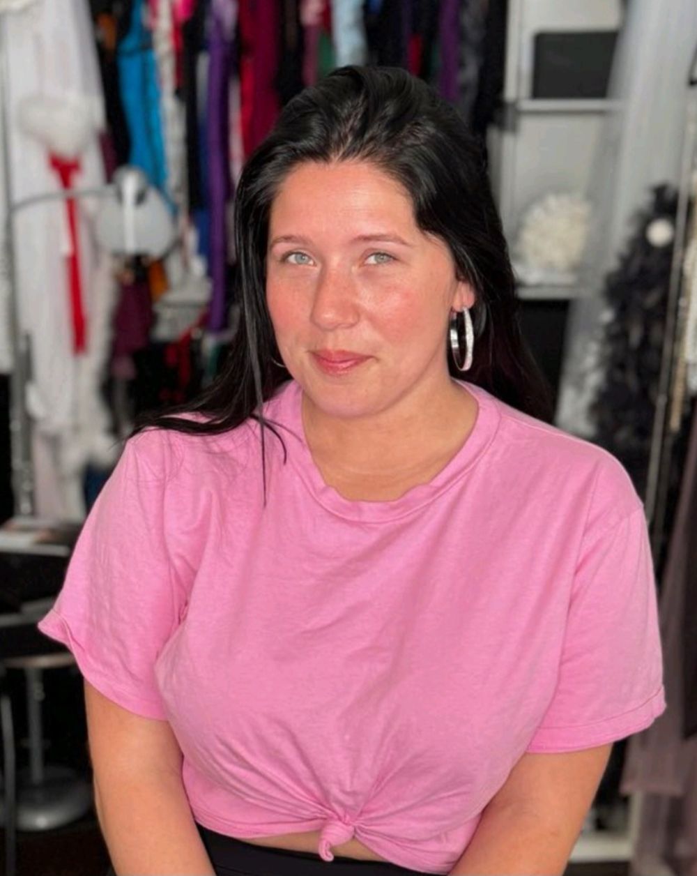 Woman in pink knotted t-shirt with hoop earrings, dark hair, standing in front of a clothing rack.
