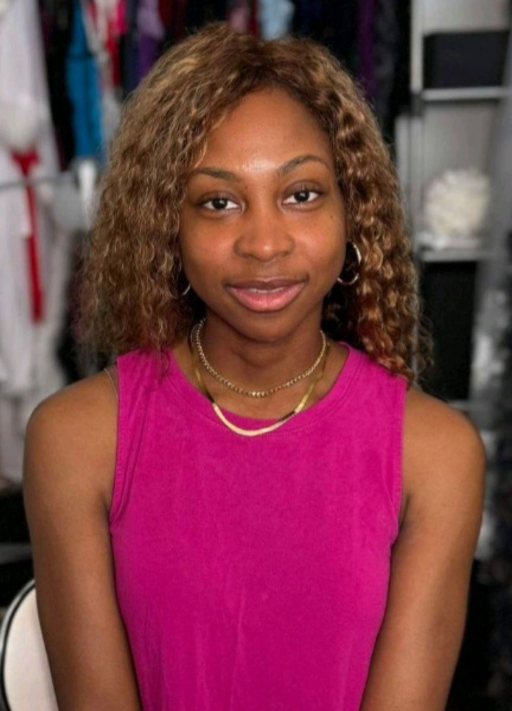 Young woman with curly hair wearing a pink sleeveless top and gold necklace, smiling in a clothing store.
