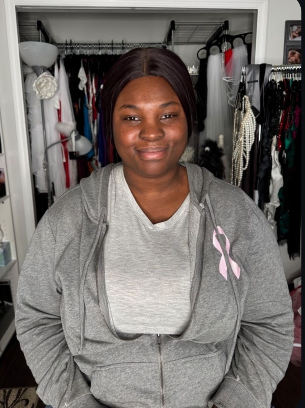 Woman in gray hoodie with pink ribbon pin smiles in front of clothing rack.