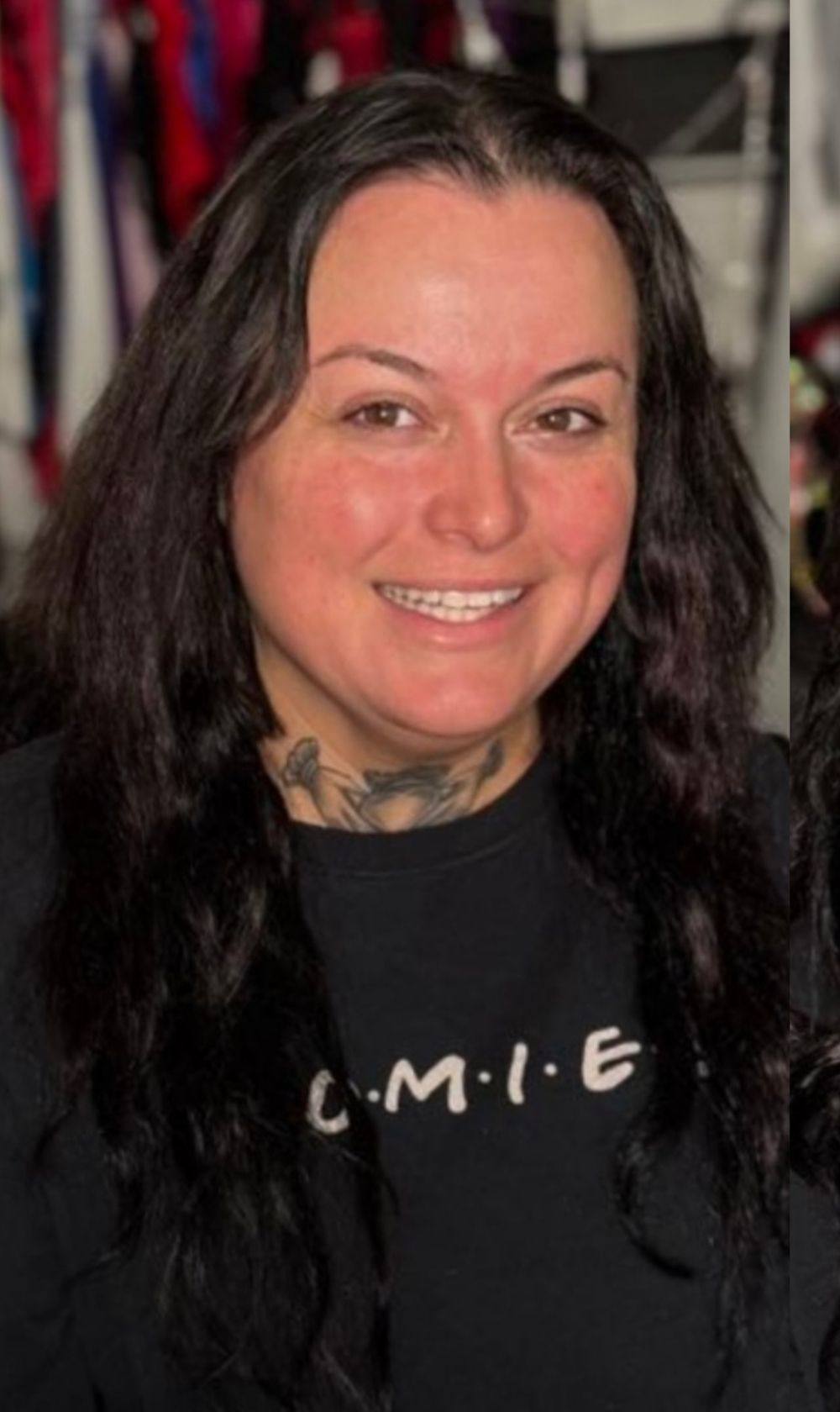 Woman with long dark hair and neck tattoo smiling, wearing a black Friends-themed sweatshirt.