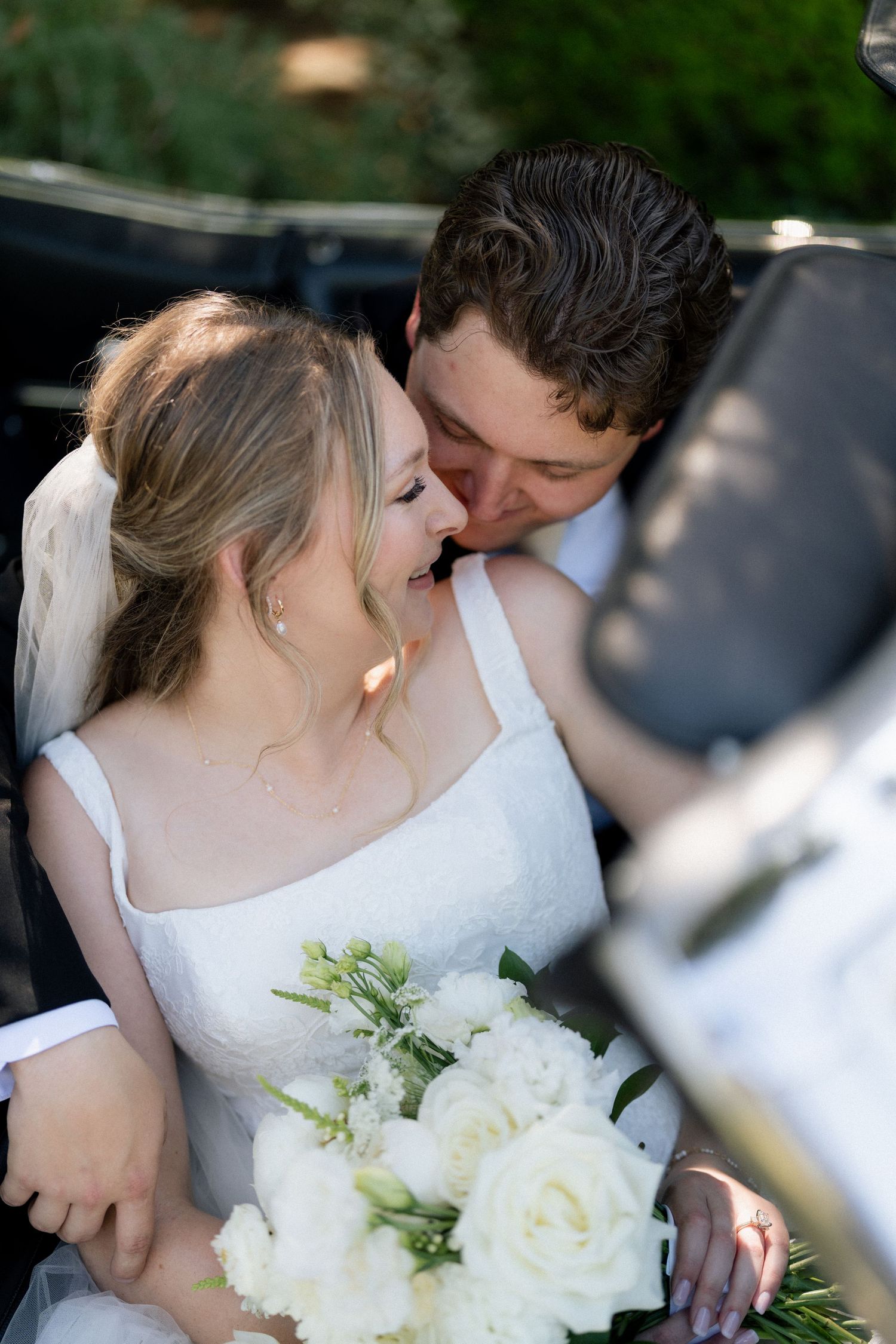 Bride in white dress holding bouquet laughs as groom kisses her cheek near a vintage car. Fresno California Wedding Photographer and Video