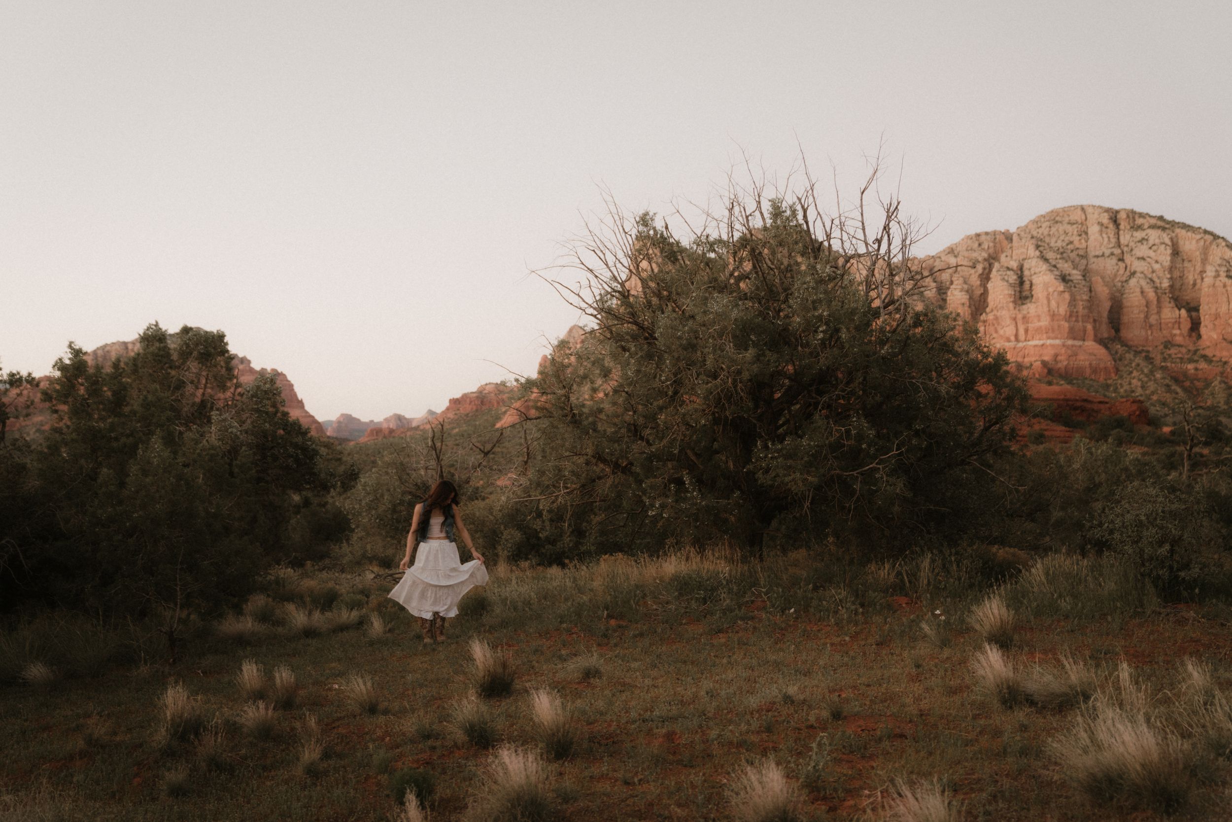 A dreamy silhouette in a white dress walks through desert brush with red cliffs in the background.