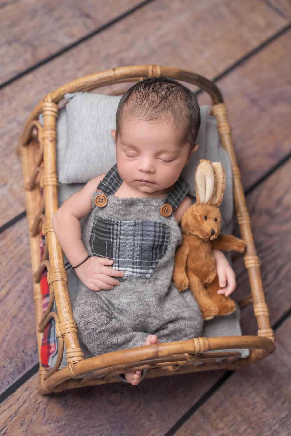 A sleeping baby in overalls rests in a wooden basket with a plush bunny toy on dark wooden floorboards.