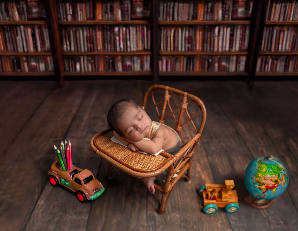 A sleeping newborn rests in a wooden chair surrounded by educational toys including a globe and toy vehicles on hardwood floor.