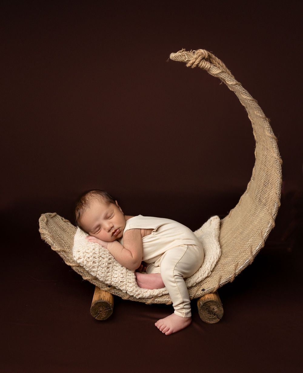 A sleeping baby lies peacefully on a rustic curved wooden piece against a dark brown background.