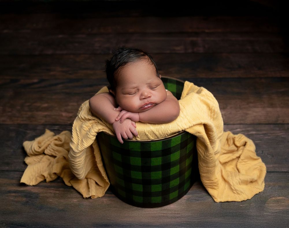 A sleeping baby nestled in a green plaid basket with soft yellow fabric on a rustic wooden background.