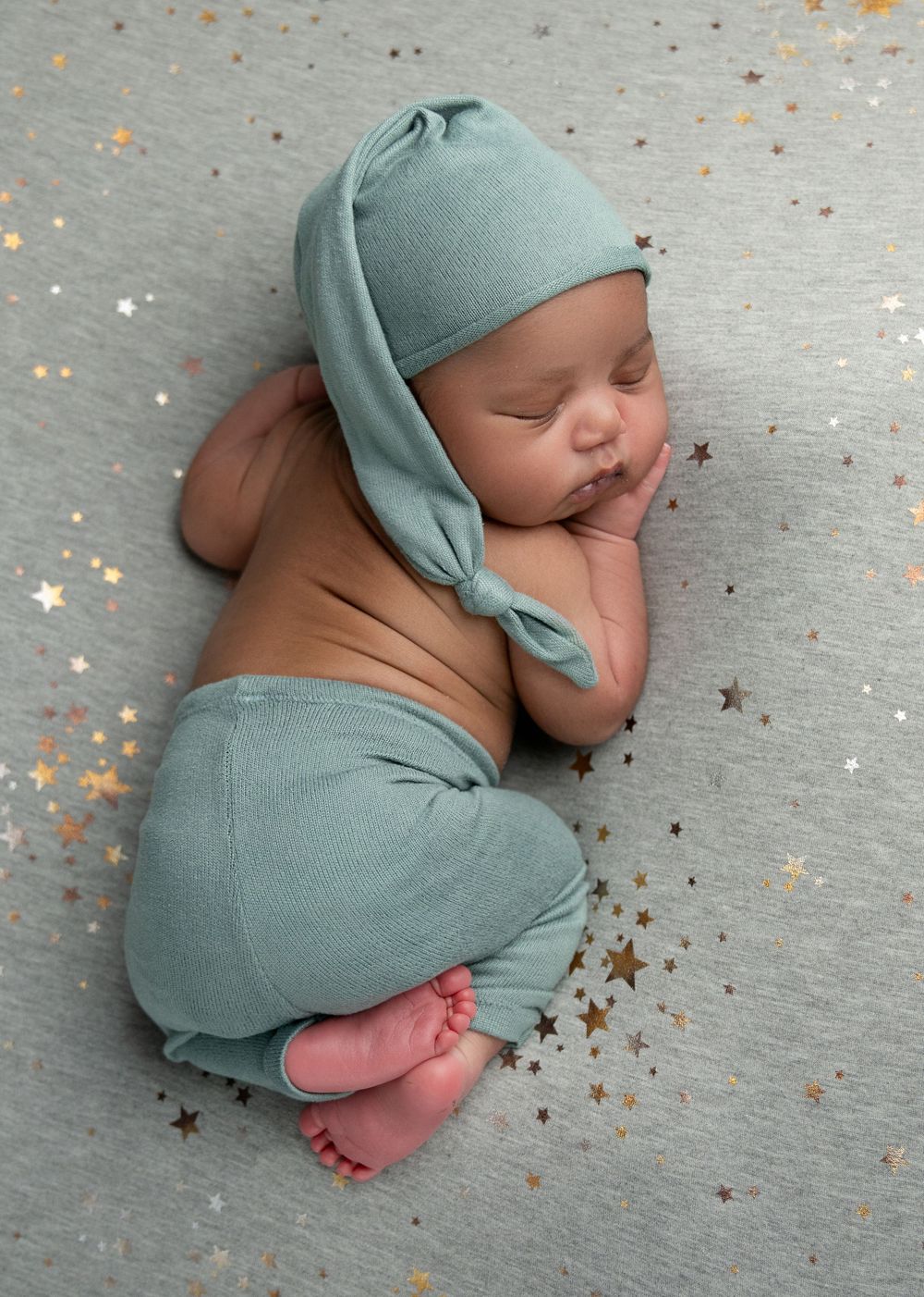 Sleeping newborn in sage green outfit lying on gray backdrop with scattered gold star confetti decorations.