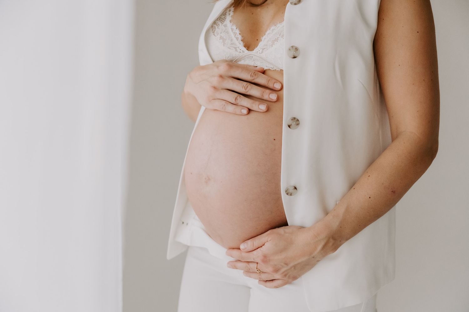Gravid kvinde i hvid kjole holder sin bare mave med begge hænder mod en lys baggrund. Fotograf Middelfart.