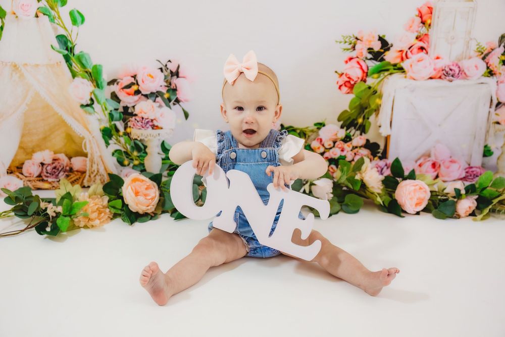 Baby girl in blue romper holds ONE sign surrounded by pink flowers for her first birthday photo session.