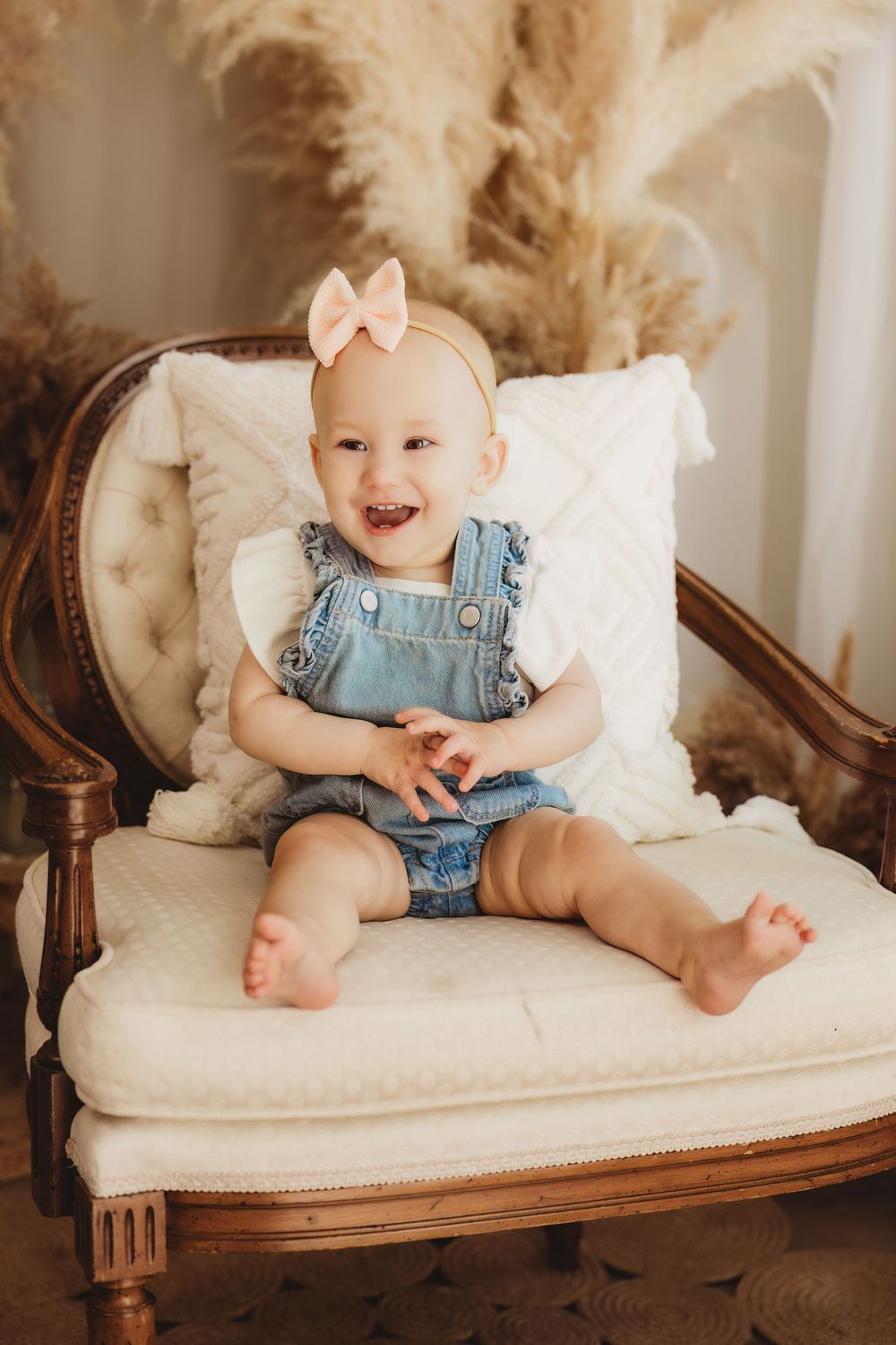 Smiling baby girl in denim overalls and pink bow headband sitting on a vintage cream armchair.