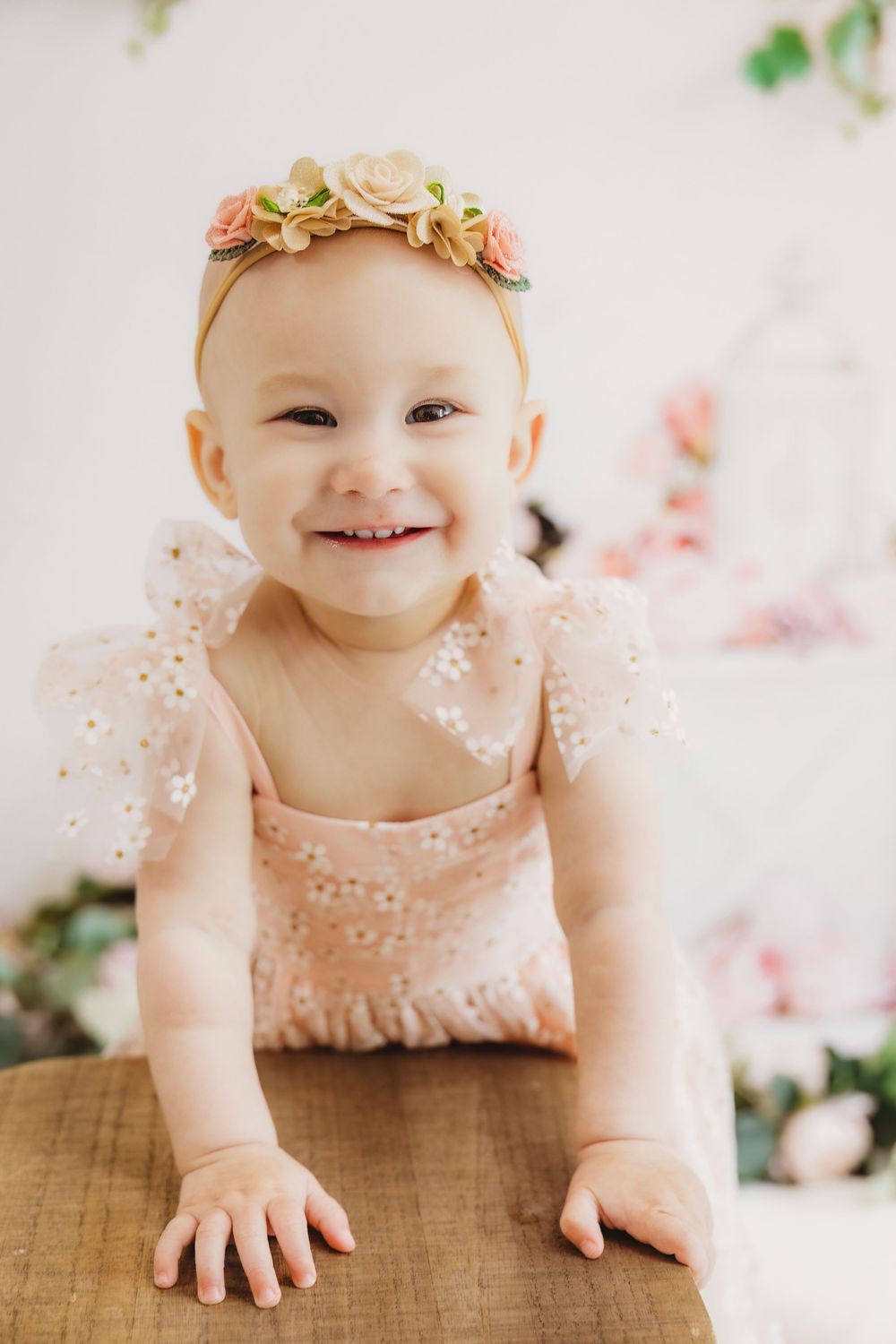 Smiling baby girl in pink floral romper and flower crown headband leaning on wooden surface in bright studio setting.