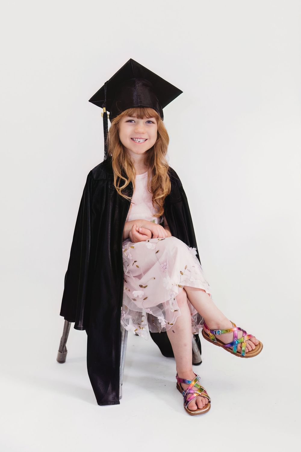 Young girl in graduation cap and gown sitting on stool, smiling, wearing floral dress and colorful sandals.