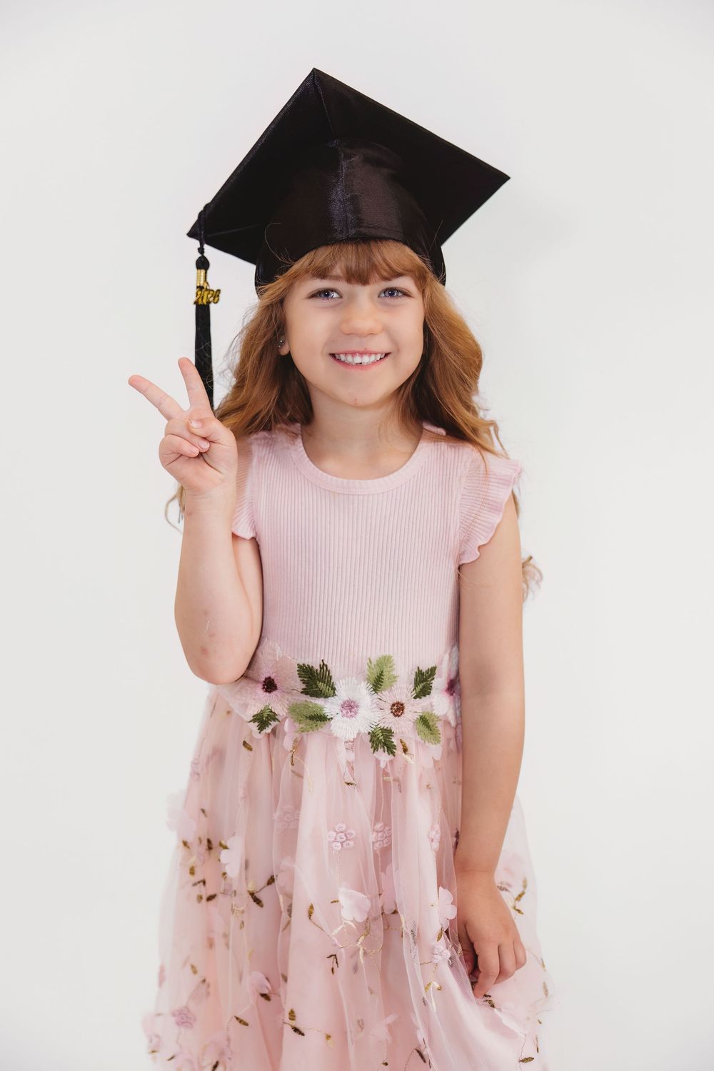 Smiling young girl in pink floral dress and graduation cap giving peace sign on white background.