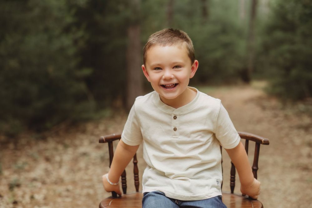 Happy young boy smiling on a wooden bench along a forest path, wearing a cream henley shirt and jeans.