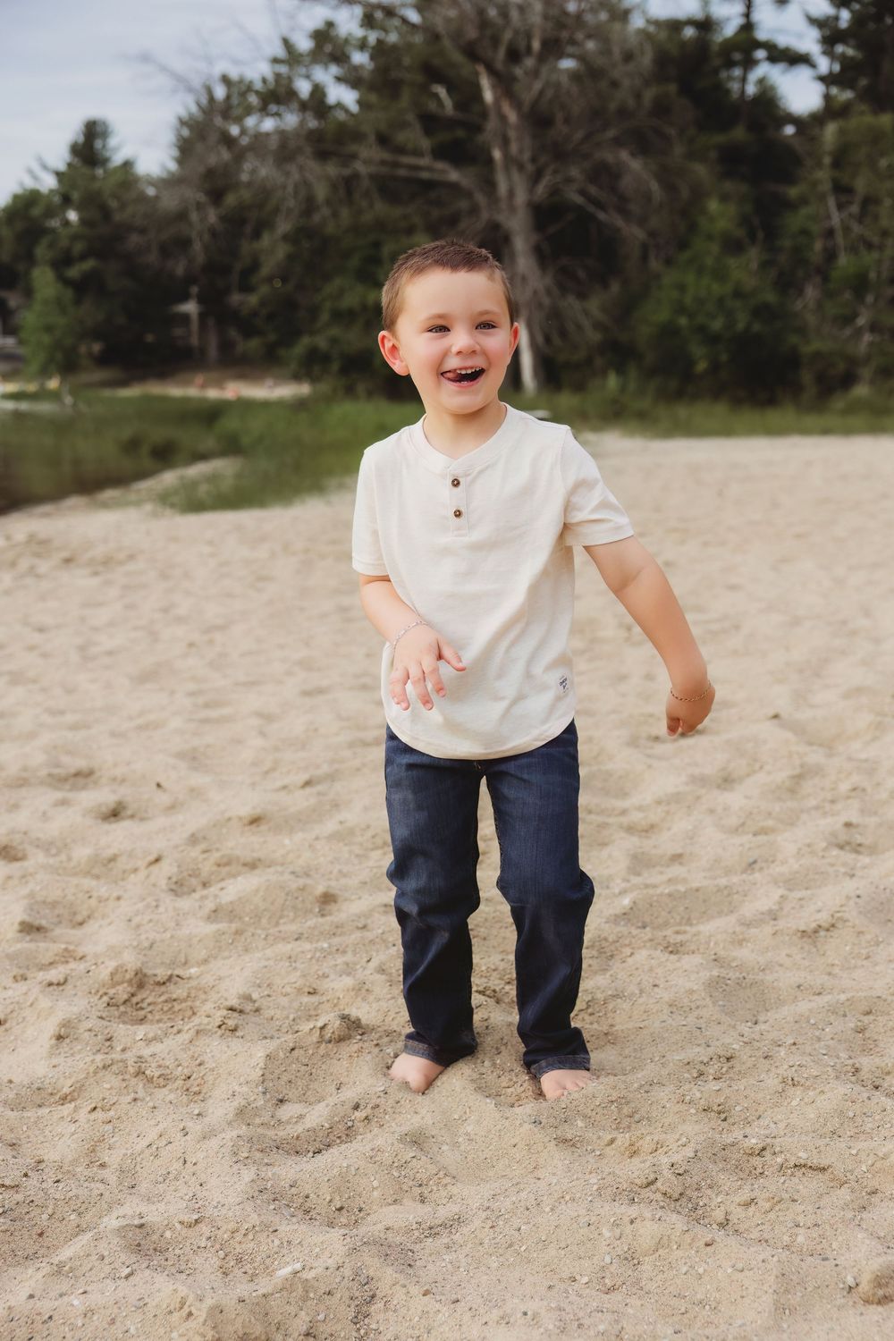 Happy young boy laughing and playing barefoot on sandy beach wearing white shirt and navy jeans.