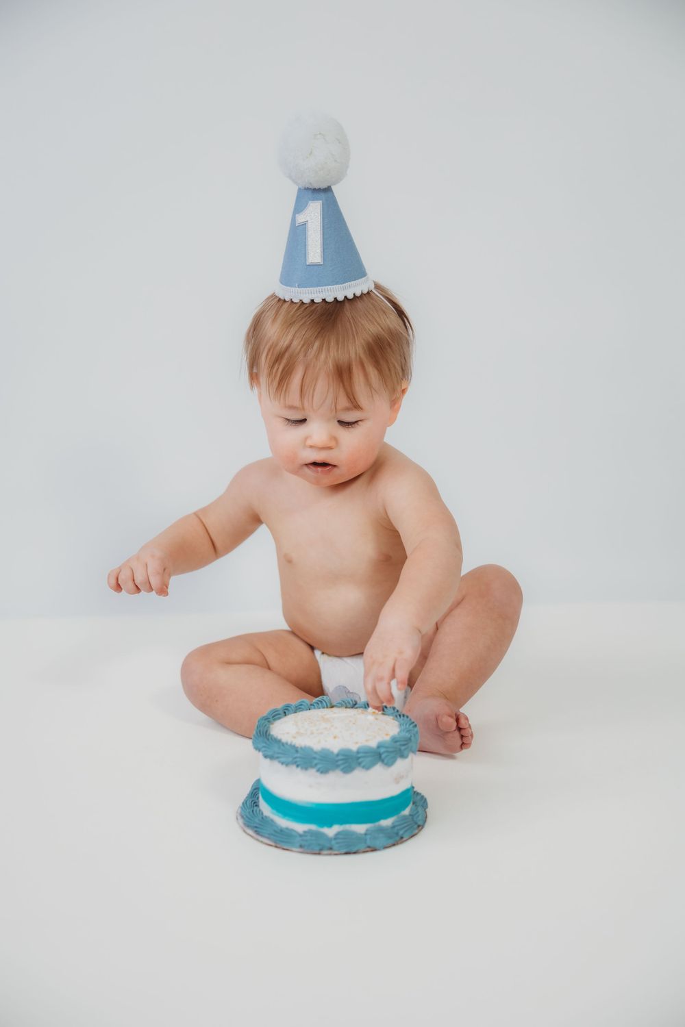 Baby wearing blue party hat reaches for birthday cake during cake smash photo session.