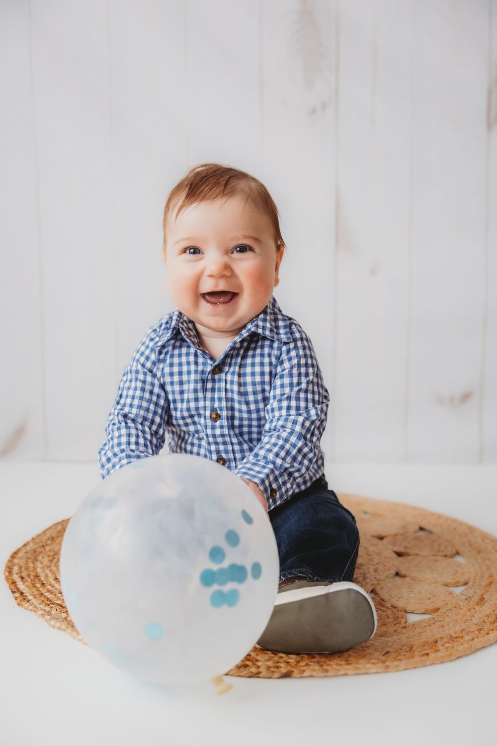 Baby wearing blue gingham shirt smiles while sitting on woven mat with decorative cloud prop.