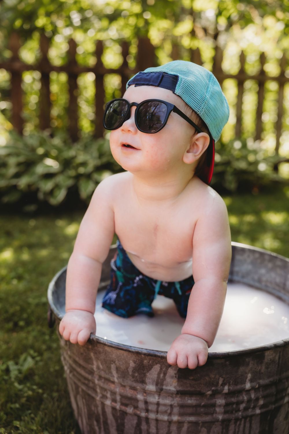 Baby wearing sunglasses and cap sits in metal basin outdoors during summer.