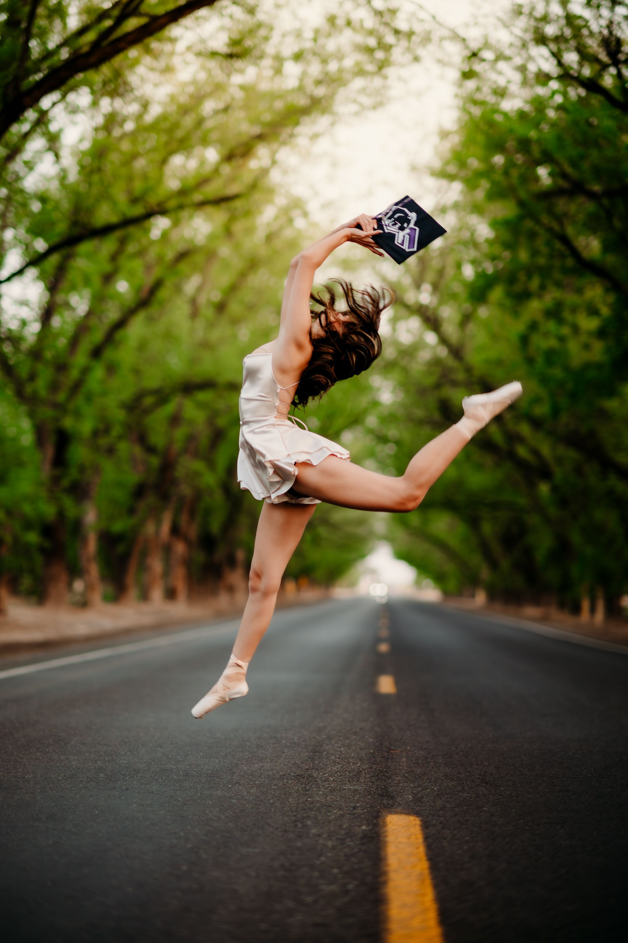Graceful ballerina performs an athletic leap on an empty tree-lined road wearing pointe shoes and a flowing white dress.