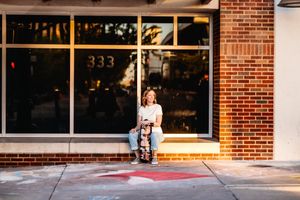 A person sits on a chair outside a storefront with large windows and brick walls in warm sunlight.