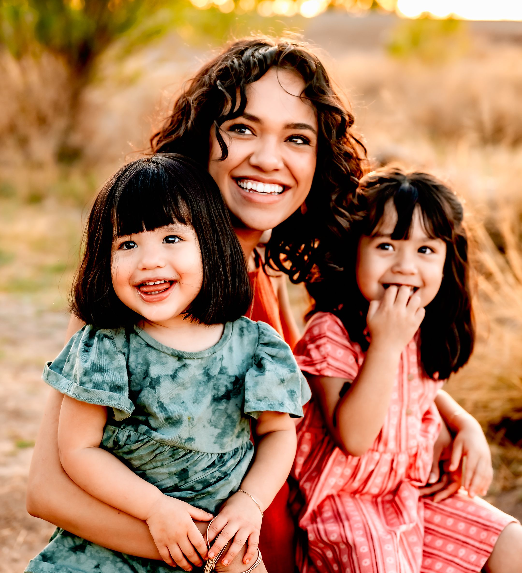 Three people sit together outdoors in casual dresses during golden hour against a natural background.