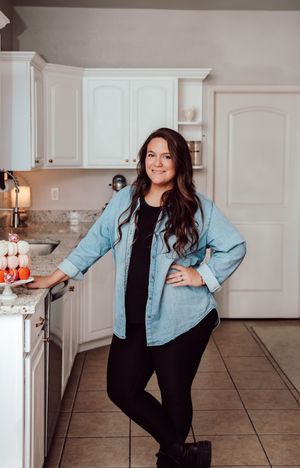 Woman in light blue denim shirt and black leggings poses casually in modern white kitchen with granite countertops.