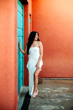 Woman in white dress poses against vibrant coral wall with turquoise door creating colorful tropical aesthetic.