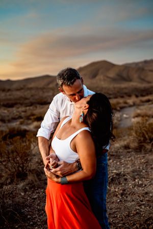 A romantic couple embraces at sunset in a desert landscape with mountains in the background.