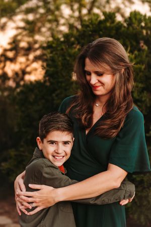 A mother in a dark green dress embraces her young son outdoors during golden hour.