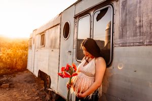 A pregnant figure in a white dress holds red flowers beside a vintage trailer during a golden sunset.