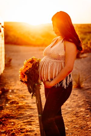 Silhouette of pregnant woman in crochet top holding sunflowers at golden sunset on dusty path.