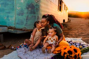 A family sits together on a blanket at sunset outside a turquoise trailer with stuffed animals nearby.