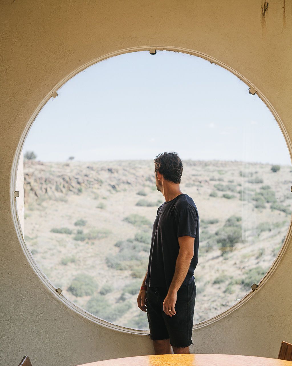 Man in black outfit stands by large circular window overlooking arid desert landscape.